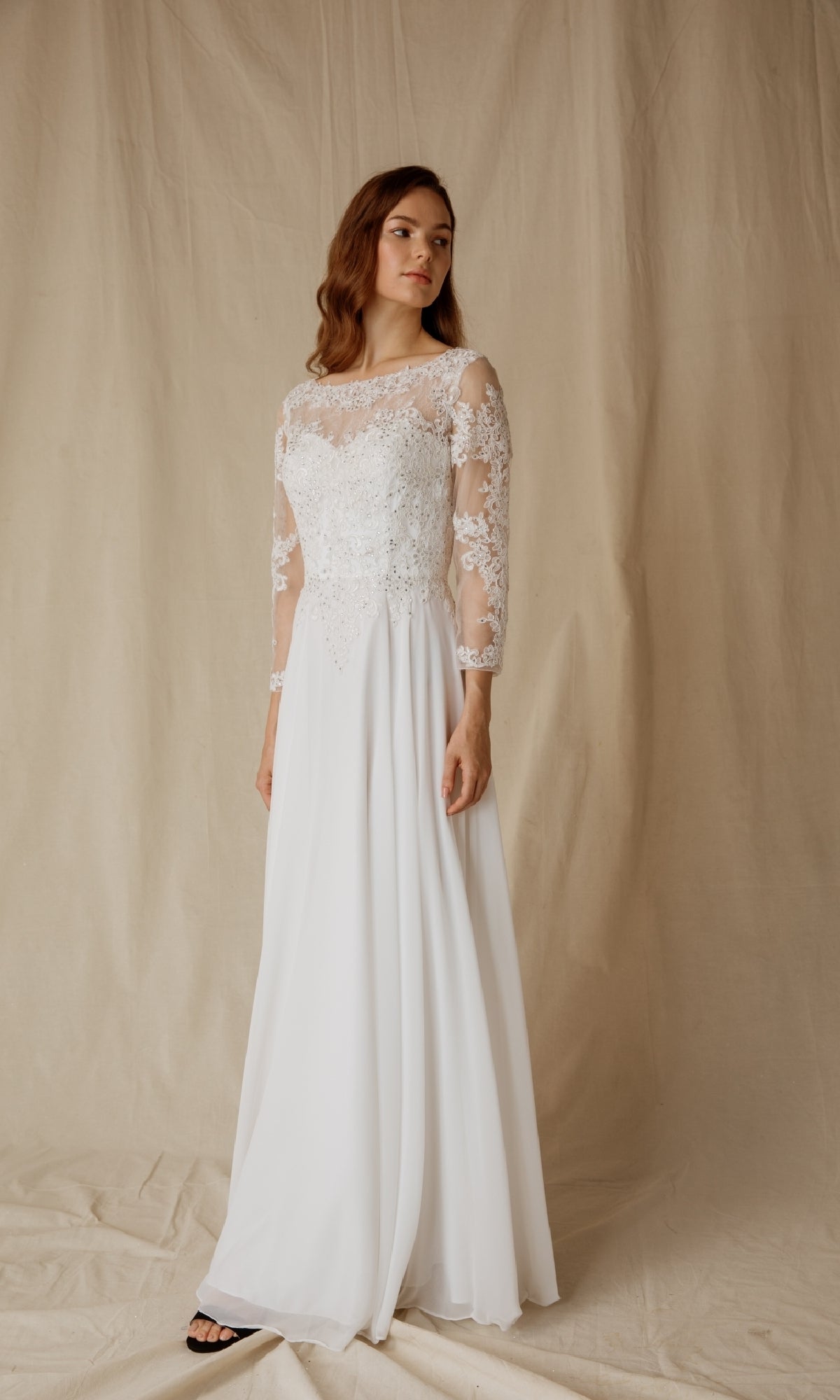 A woman in the Eureka 6909 long-sleeve formal dress with a lace bodice stands on a light fabric backdrop, gazing to the side with a neutral expression.